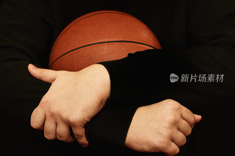 穿着深色毛衣的篮球运动员er<e:2>的手在暖橙色的灯光下抱着一个黑色背景的篮球，篮球在黑色背景中